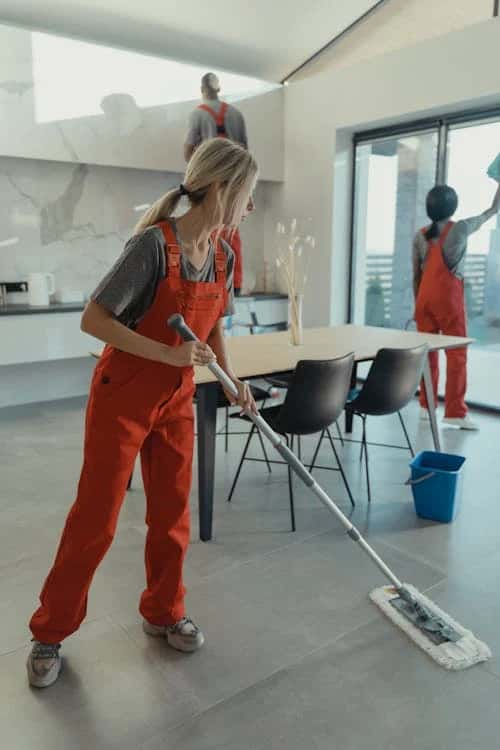 A woman in overalls cleaning a room. She is holding a mop and a bucket, focused on tidying up the space.