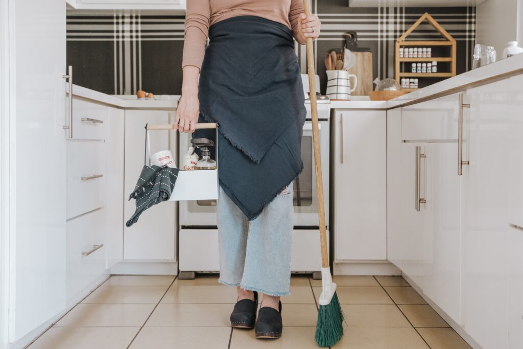 Woman standing in a kitchen carrying a basket of cleaning supplies.