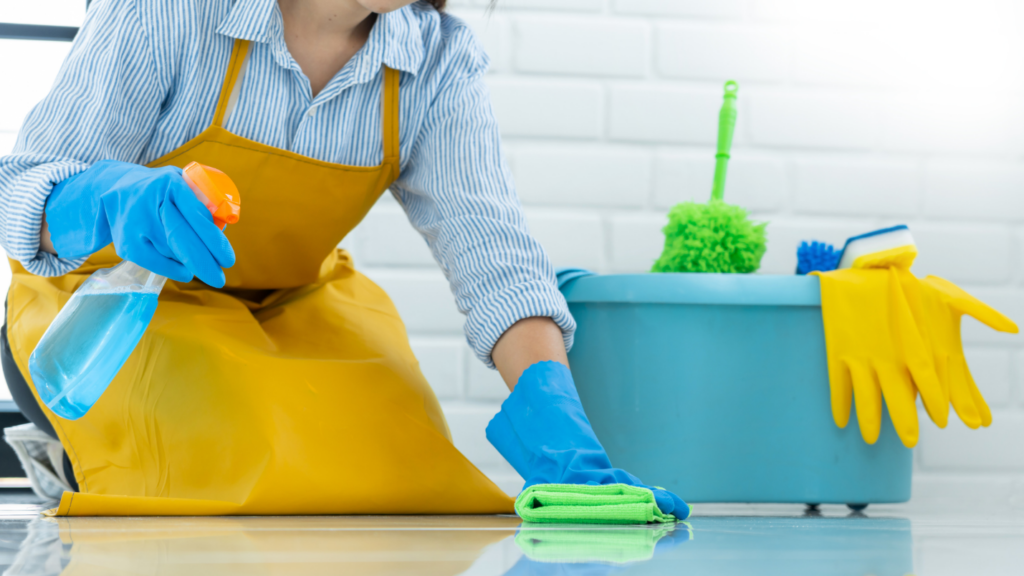 A maid in an apron holds a bottle of cleaning spray and scrubs the floor with a towel.