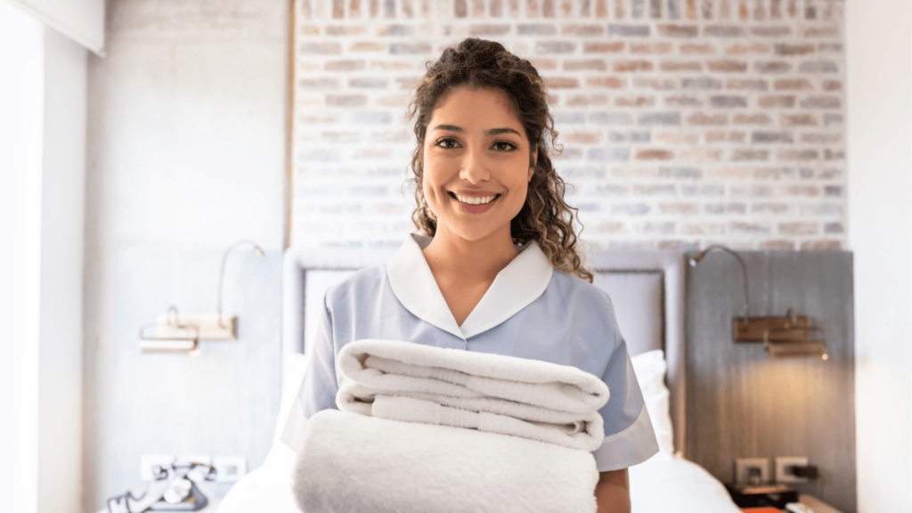 A maid wearing a light blue uniform smiles while holding freshly folded towels.