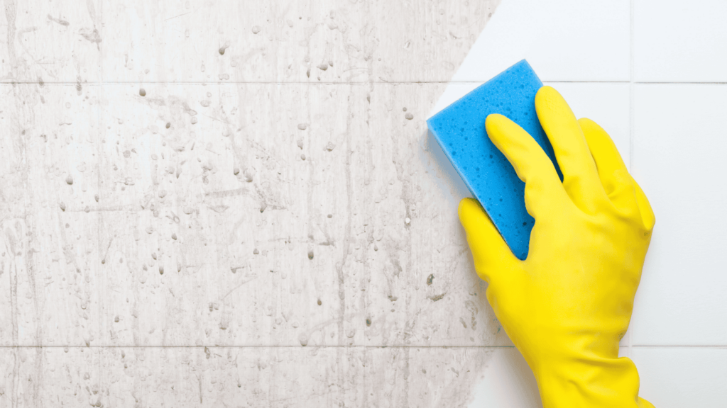 A hand wearing a rubber glove uses a sponge to clean a tile wall.